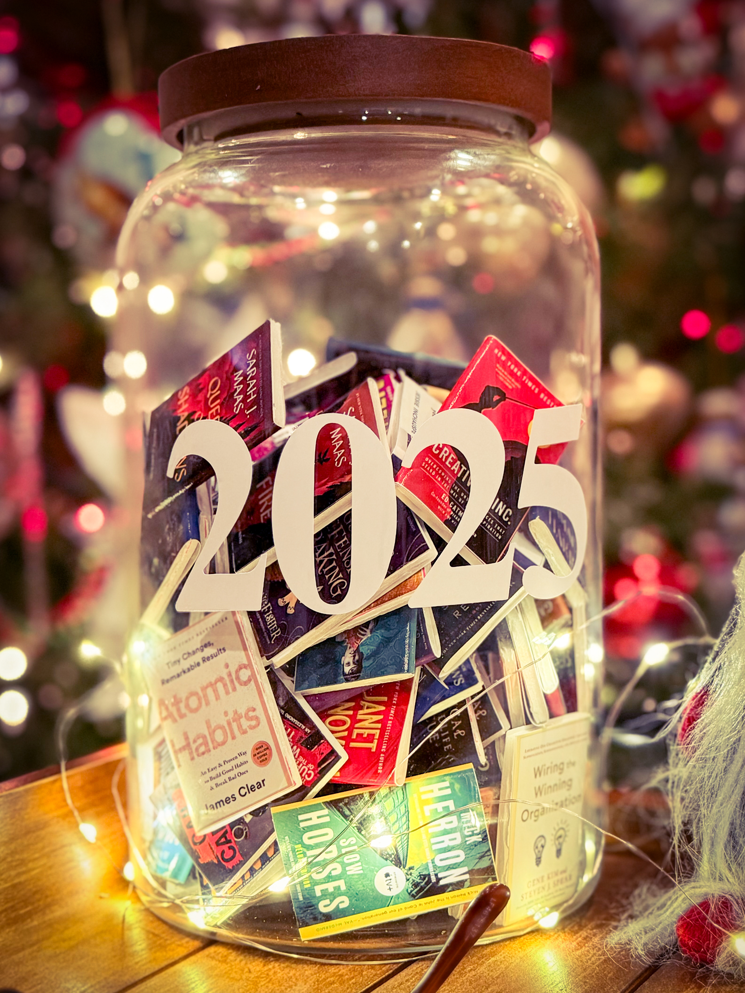 A large glass jar filled with hand-cut miniature book covers, topped with a paper 2025 cutout, lit by fairy lights in front of a holiday tree
